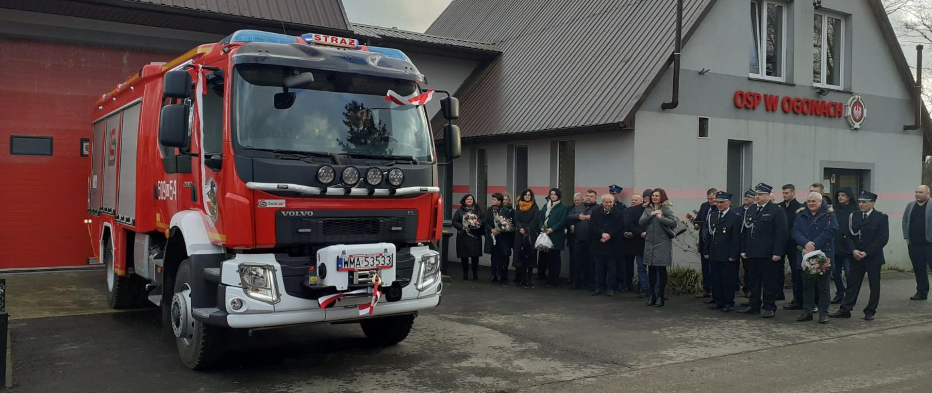 Strażacy oraz zaproszeni goście stoją wokół samochodu pożarniczego na placu przed remizą.