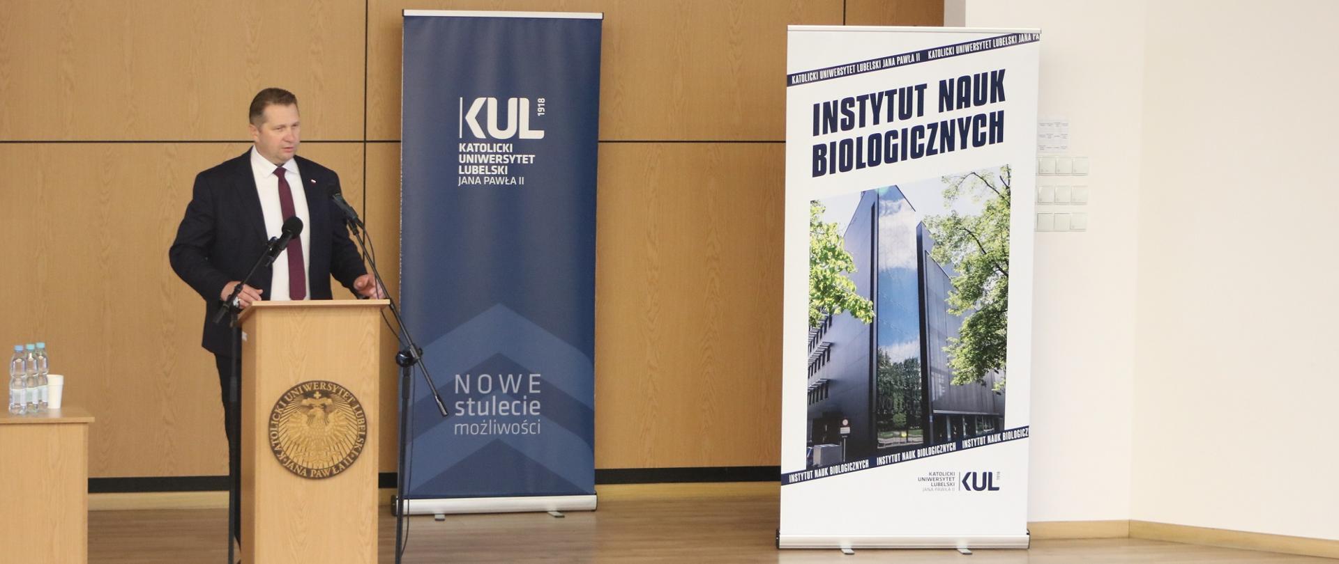Minister Czarnek stoi przy mównicy i mówi do mikrofonu, za nim brązowa ściana, przy niej plakat z napisem KUL i Instytut Nauk Biologicznych.