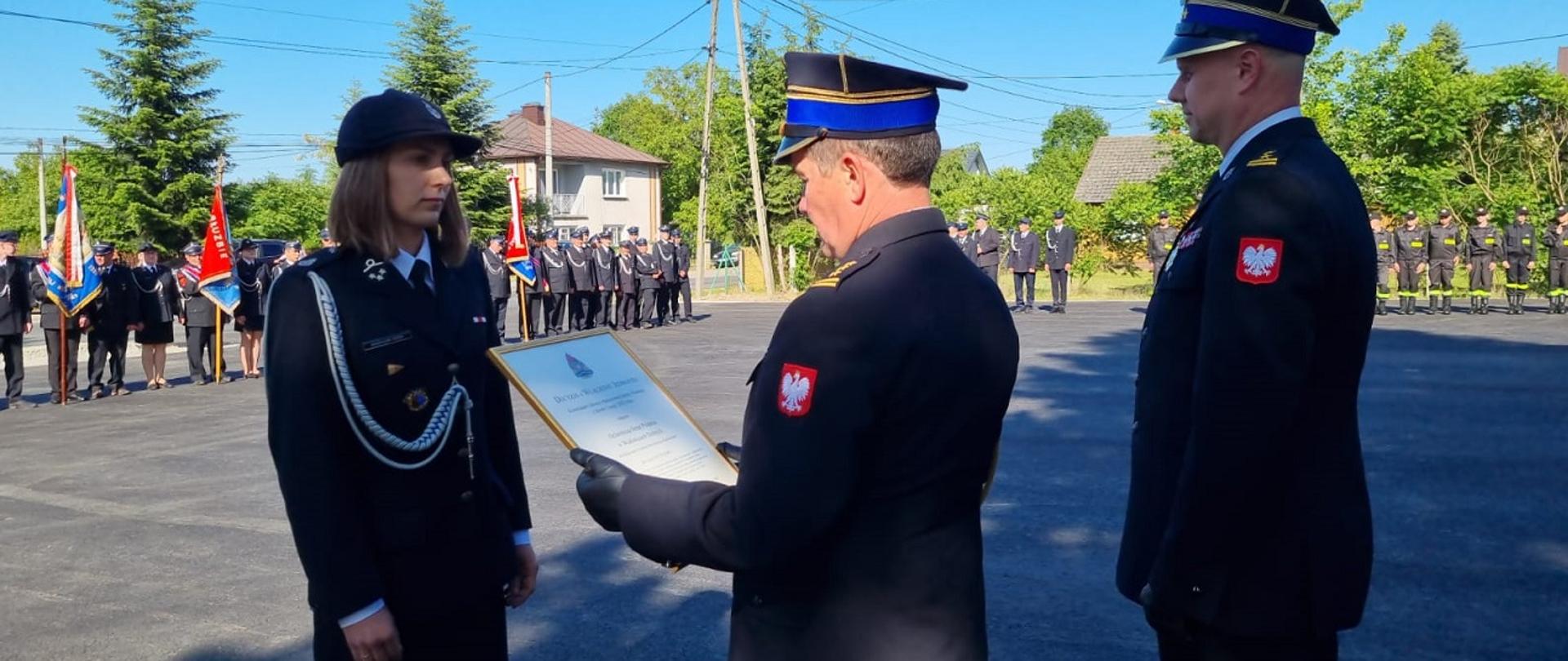 Fotografia wykonana podczas uroczystości gminnych obchodów dnia strażaka i włączenia jednostki ochotniczej straży pożarnej do krajowego systemu ratowniczo-gaśniczego. Na zdjęciu zastępca podkarpackiego komendanta wojewódzkiego Państwowej Straży Pożarnej w asyście komendanta powiatowego Państwowej Straży Pożarnej w Mielcu, wręcza druhnie która jest członkiem jednostki Ochotniczej Straży Pożarnej w Wadowicach Dolnych , decyzję o włączeniu jednostki do krajowego systemu ratowniczo-gaśniczego. Funkcjonariusze i druhna ubrani w mundury galowe koloru granatowego stoją na środku placu uroczystej akademii. W tle widać poczty sztandarowe oraz strażaków ochotników stojących wokół placu.