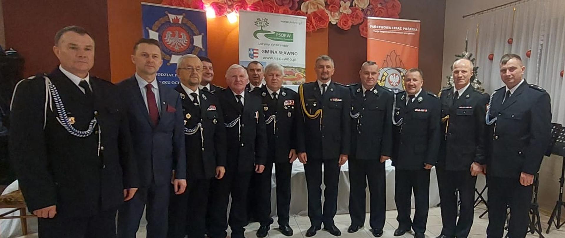 Zdjęcie z sali. W pozycji stojącej w umundurowaniu Komendanci PSP i druhowie OSP oraz przedstawiciele samorządu, łącznie 12 osób. W tle logo ochotniczej straży pożarnej, logo gminy Sławno oraz logo Państwowej Straży Pożarnej. 