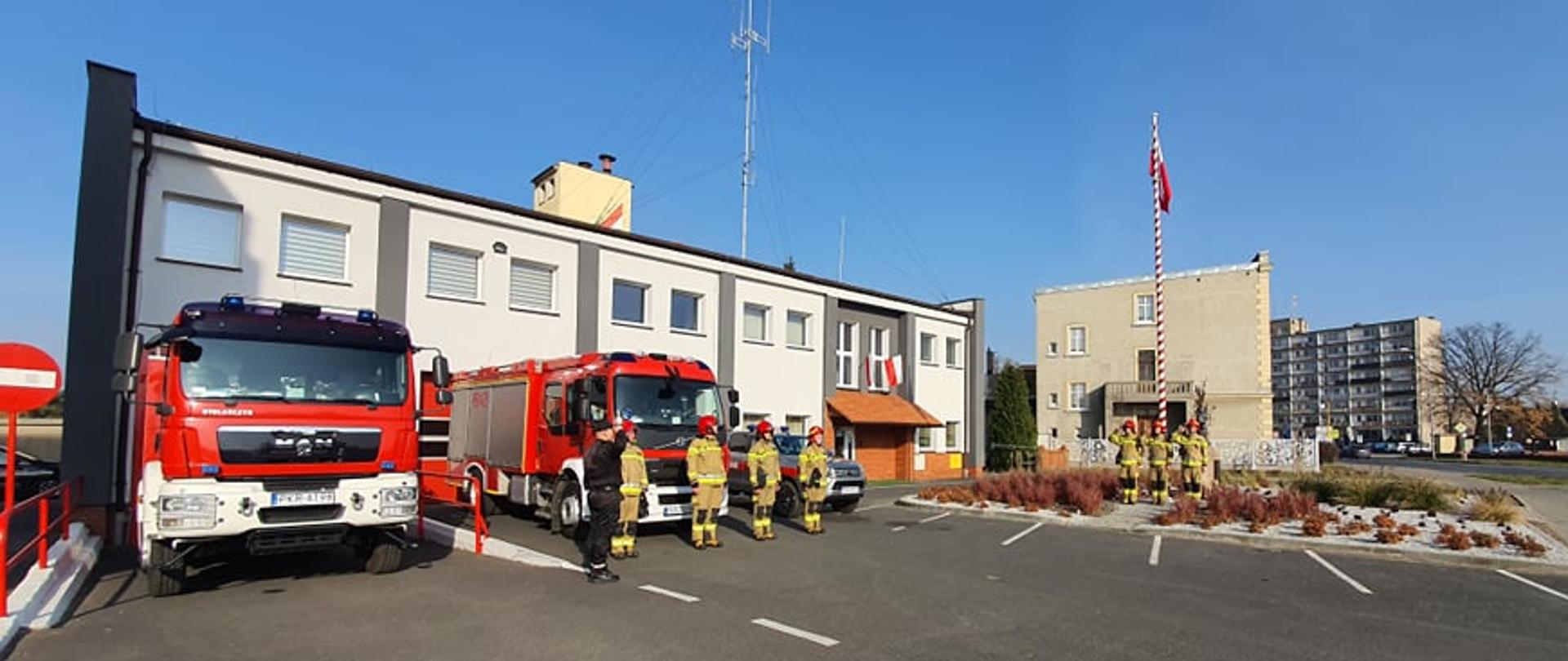 Strażacy oddają honor podniesionej na maszt fladze państwowej w tle trzy samochody pożarnicze przed budynkiem KP PSP w Krotoszynie