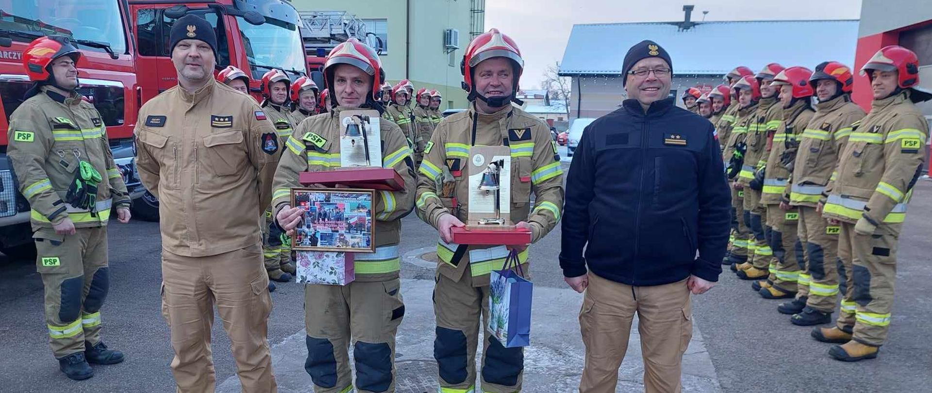 Funkcjonariusze odchodzący na zaopatrzenie emerytalne trzymają w rękach upominki. Obok nich stoją Komendancji. Za nimi na placu komendy stoją zmiany służbowe.