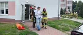 Na zdjęciu widoczni są strażak, który ewakuuje mężczyzn z budynku szpitala i kieruj się w miejsce ewakuacji. W tle widoczni są strażacy, którzy kierują się realizować zadania wyznaczone przez Kierującego Działaniami Ratowniczymi.