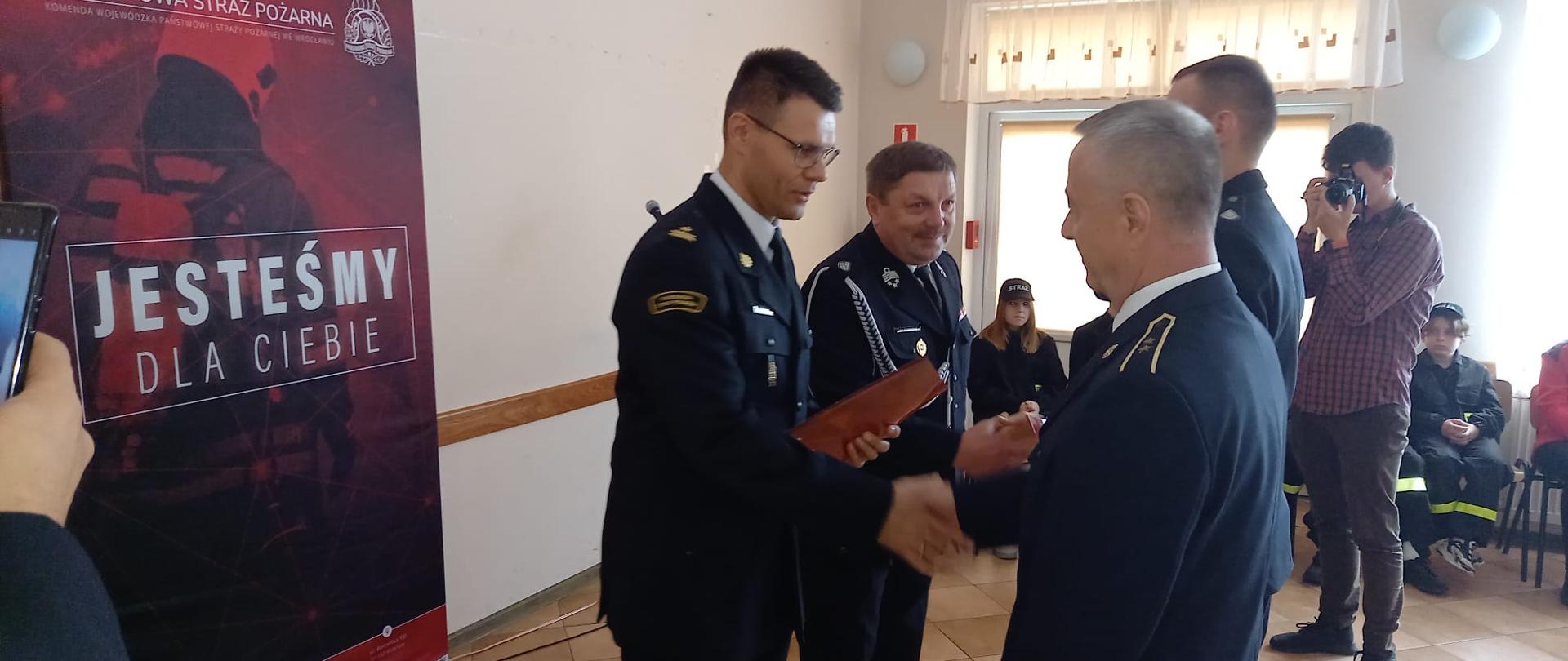 Obraz przedstawia Zastępcę Komendanta Powiatowego wręczającego promesy dla MDP. Osoby w umundurowaniu wyjściowym w świetlicy wiejskiej.