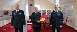 Na zdjęciu znajdują się trzej strażacy w umundurowaniu galowym. W środku znajduj się strażak przechodzący na zaopatrzenie emerytalne, który trzyma w rękach grawerton gratulacyjny. Strażacy umiejscowieni są w pomieszczeniu sali tradycji Komendy Powiatowej Państwowej Straży Pożarnej w Jędrzejowie. W tle widnieją tablice z ważnymi wydarzeniami historycznymi Komendy Powiatowej PSP w Jędrzejowie. 