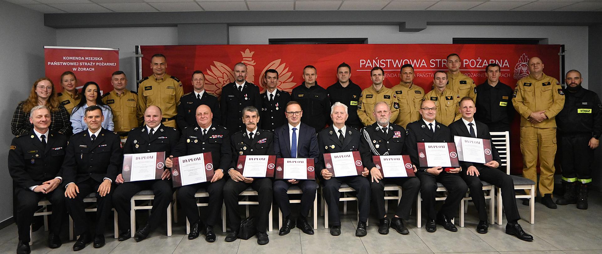 Strażacy w mundurach ustawieni na tle czerownego baneru. W pierwszym rzędzie trzymają dyplomy i siedzą na krzesłach. Pomiędzy nimi stoi kilka osób cywlnych. Nad ich głowami napis Państwowa Straż Pożarna.