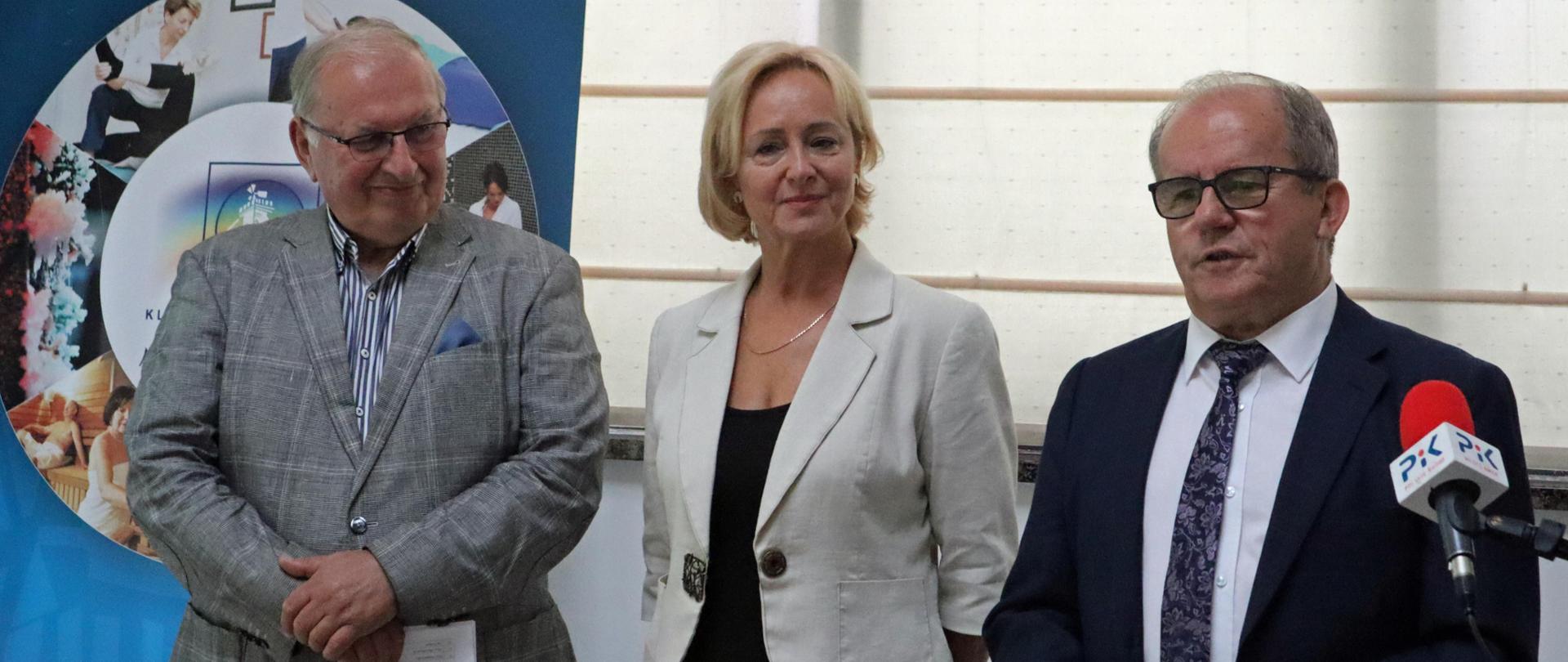 Dwóch mężczyzn i jedna kobieta stoją. Mężczyzna z prawej przemawia. 