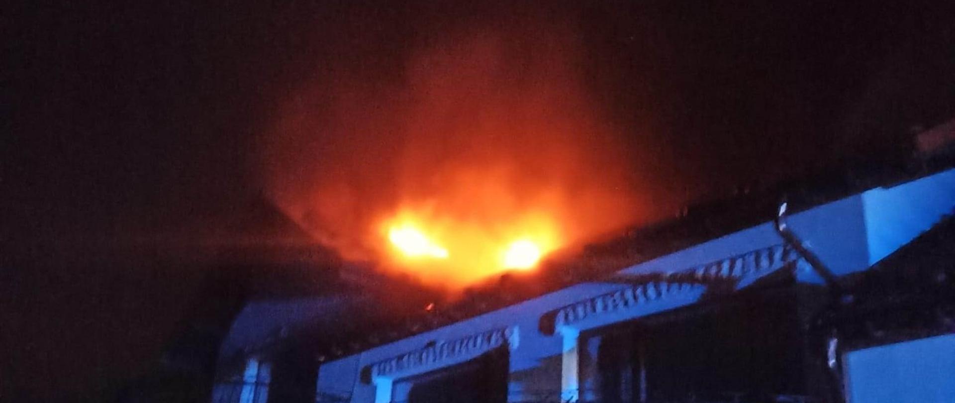 Pożar dachu na budynku mieszkalnym. Przed budynkiem stoi dwóch mężczyzn oraz 3 strażaków rozwijających linię gaśniczą. 