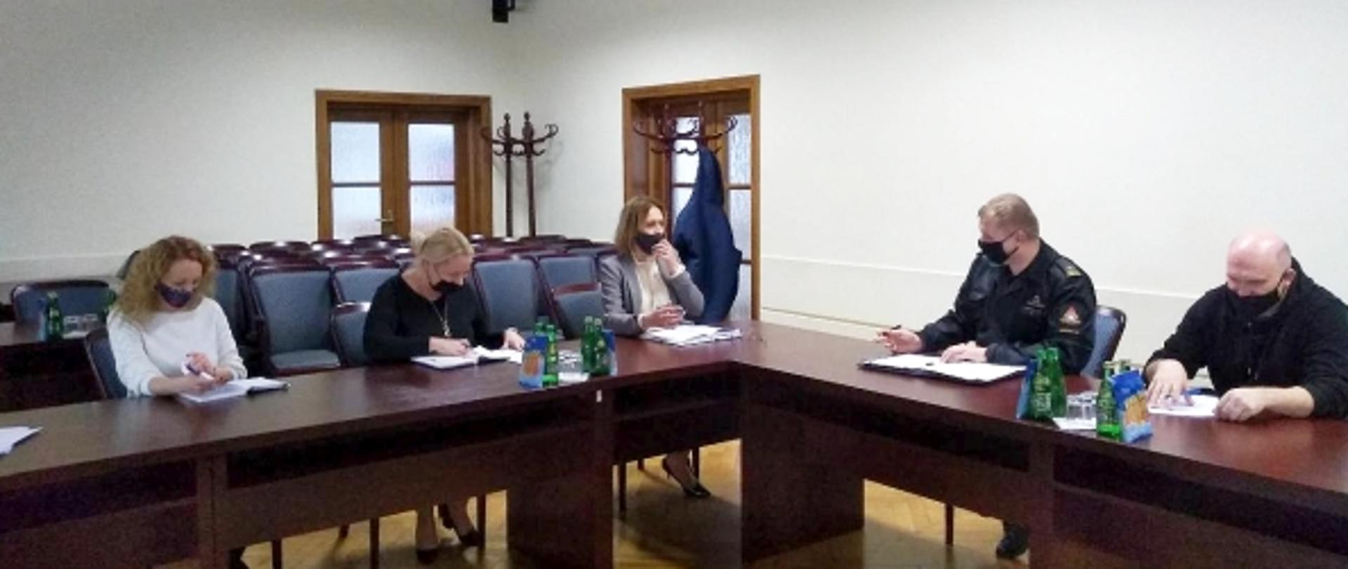 sala konferencyjna w Gminie Kozienice, 5 osób przy stole: Zastępca Burmistrza Gminy Kozienice Pani Dorota Stępień, Komendant Powiatowy Państwowej Straży pożarnej w Kozienicach mł. bryg. Kamil Bieńkowski, Komendant Gminny w Kozienicach i dwie przedstawicielki gminy Kozienice zajmujące się akcją szczepień.