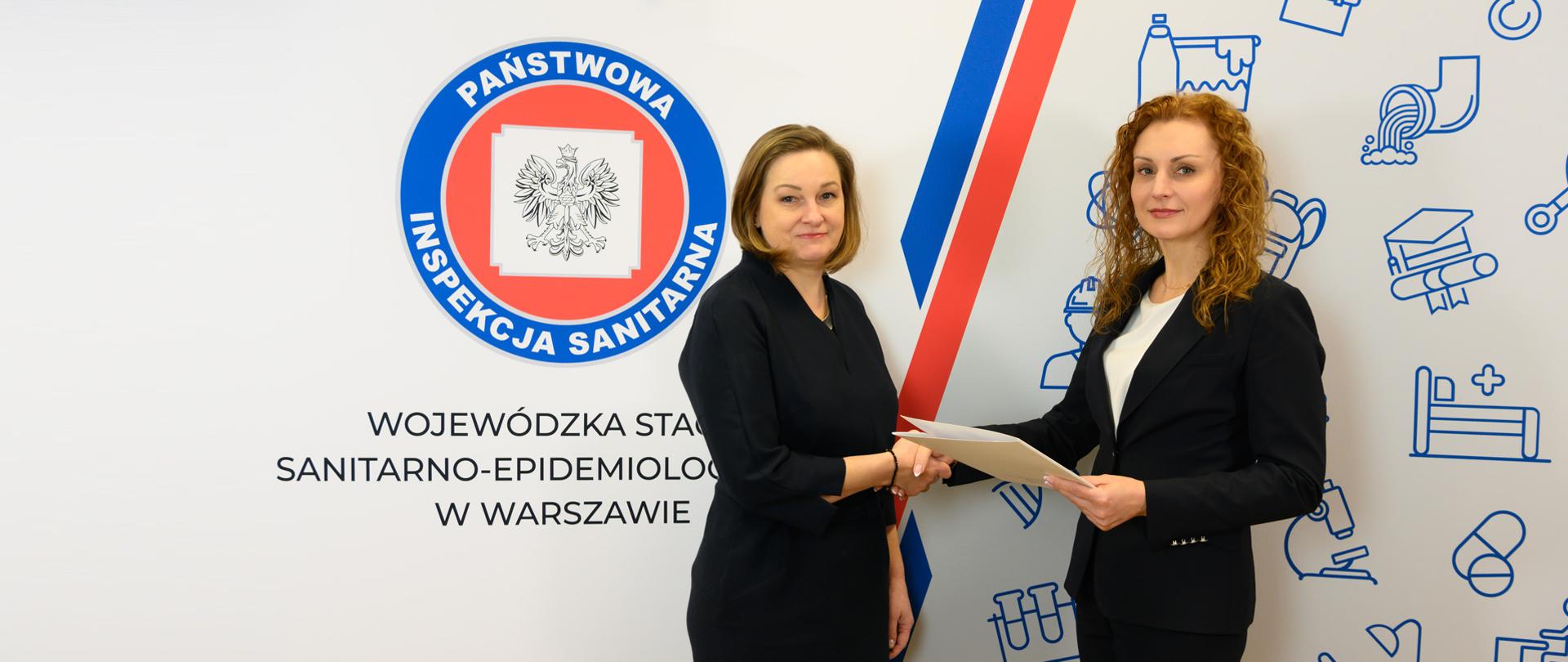 2 kobiety ściskają sobie ręce. Jedna wręcza teczkę z powołaniem. W tle logo PIS.