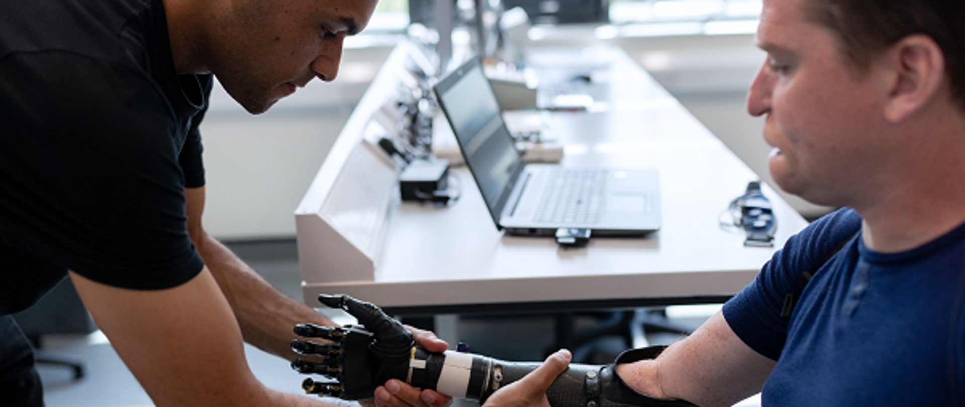 inżynier, naukowiec pracujący nad protezą prawej ręki i dłoni u siedzącego pacjenta. Proteza jest elektroniczna, nałożona od zgięcia łokciowego. W tle obu panów widać komputer stojący na blacie biurka