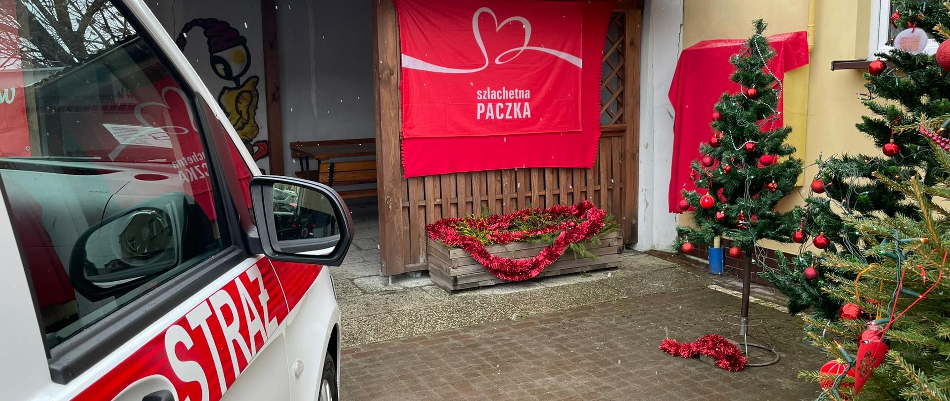 Zdjęcie przedstawia samochód pożarniczy typu bus oraz plakat na którym widnieje napis Szlachetna paczka. Dookoła widoczne choinki przystrojone w czerwone bombki. 