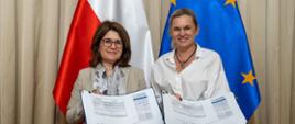 Dwie uśmiechnięte kobiety stojące obok siebie na tle flag Polski i Unii Europejskiej. Obie trzymają otwarte teczki z podpisanymi planami pracy, prezentując je do zdjęcia. Po lewej przedstawicielka UNICEF, po prawej przedstawicielka MEN .