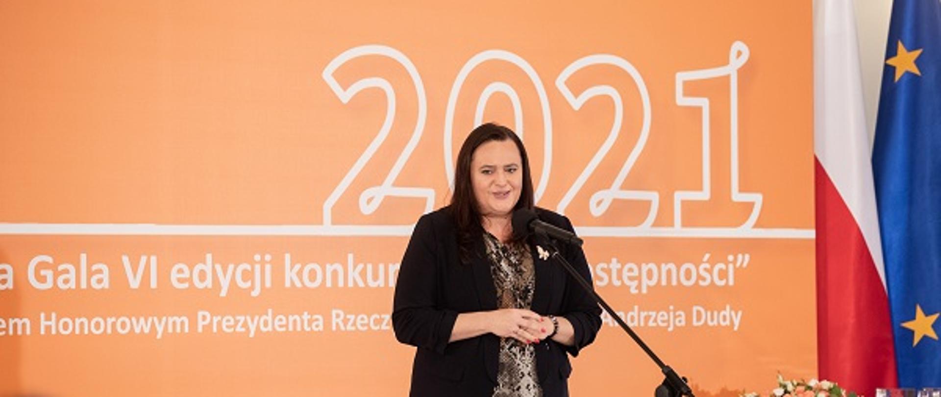 Wiceminister Małgorzata Jarosińska-Jedynak stoi na scenie przed mikrofonem. W tle napis: 2021, Gala Vi edycji konkursu Lider Dostępności, pod honorowym patronatem Prezydenta RP Andrzeja Dudy.
