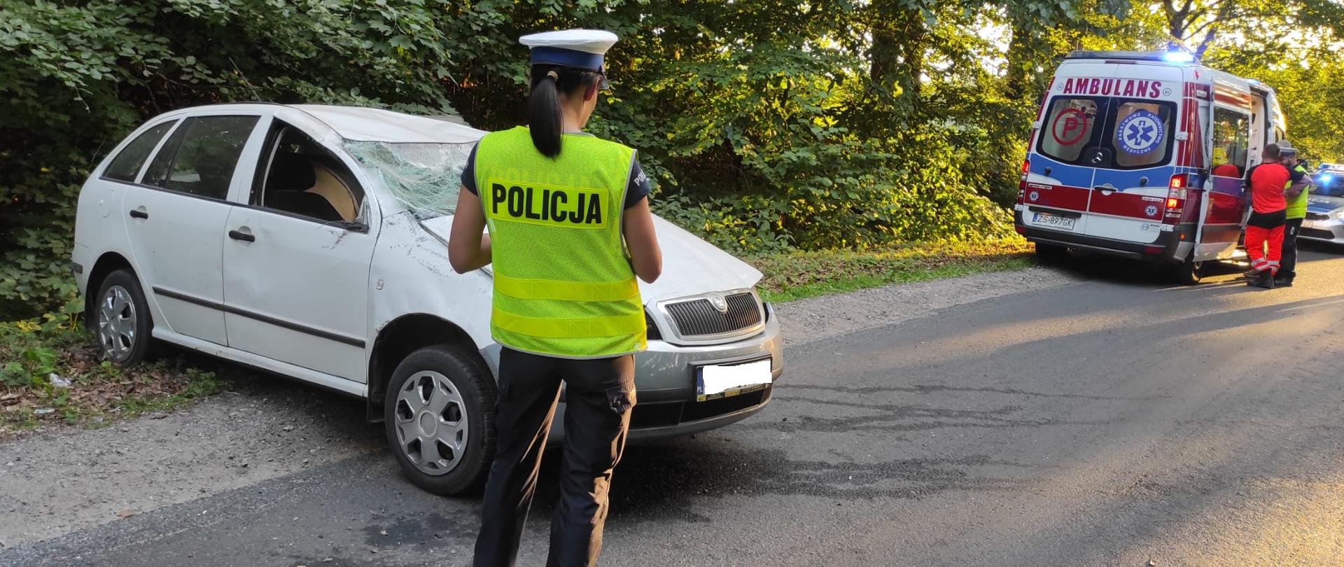 Zdjęcie przedstawia biały samochód po dachowaniu, policjantka stoi przed nim w tle karetka