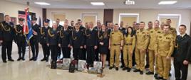 Grupowe zdjęcie uczestników uroczystości w KP PSP w Przasnyszu; strażacy w mundurach wyjściowych i koszarowych, przed nimi pamiątki