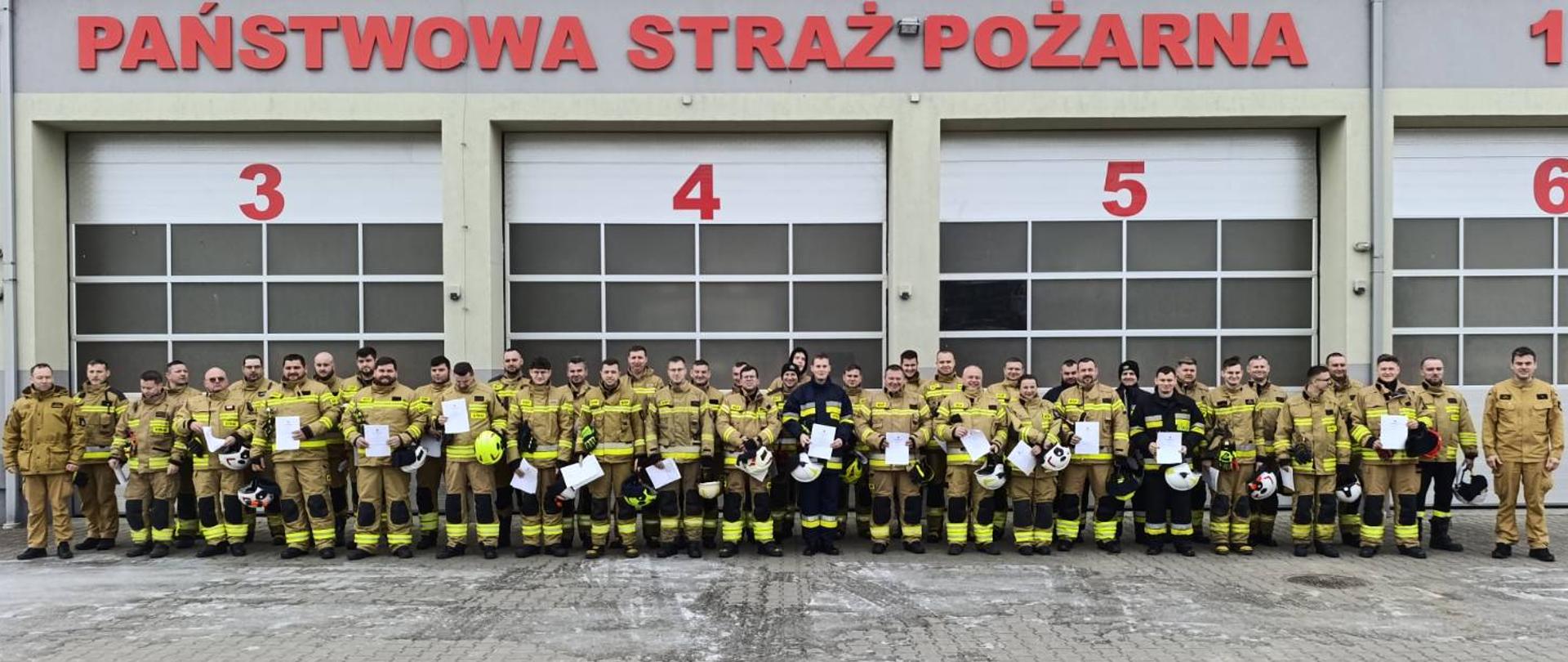 Zdjęcie przedstawia uczestników szkolenia kierowcy konserwatora. W dwuszeregu stoją strażacy w ubraniach specjalnych koloru piaskowego i granatowego. W rękach trzymają zaświadczenia o ukończeniu szkolenia koloru białego oraz kaski koloru białego i żółtego. W tle garaż JRG w Sokołowie Podlaskim koloru szarego z widocznym napisem Państwowa Straż Pożarna koloru czerwonego. Pozujący stoją na placu wyłożonym szarą kostką z elementami lodu na niej.