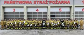 Zdjęcie przedstawia uczestników szkolenia kierowcy konserwatora. W dwuszeregu stoją strażacy w ubraniach specjalnych koloru piaskowego i granatowego. W rękach trzymają zaświadczenia o ukończeniu szkolenia koloru białego oraz kaski koloru białego i żółtego. W tle garaż JRG w Sokołowie Podlaskim koloru szarego z widocznym napisem Państwowa Straż Pożarna koloru czerwonego. Pozujący stoją na placu wyłożonym szarą kostką z elemantami lodu na niej.