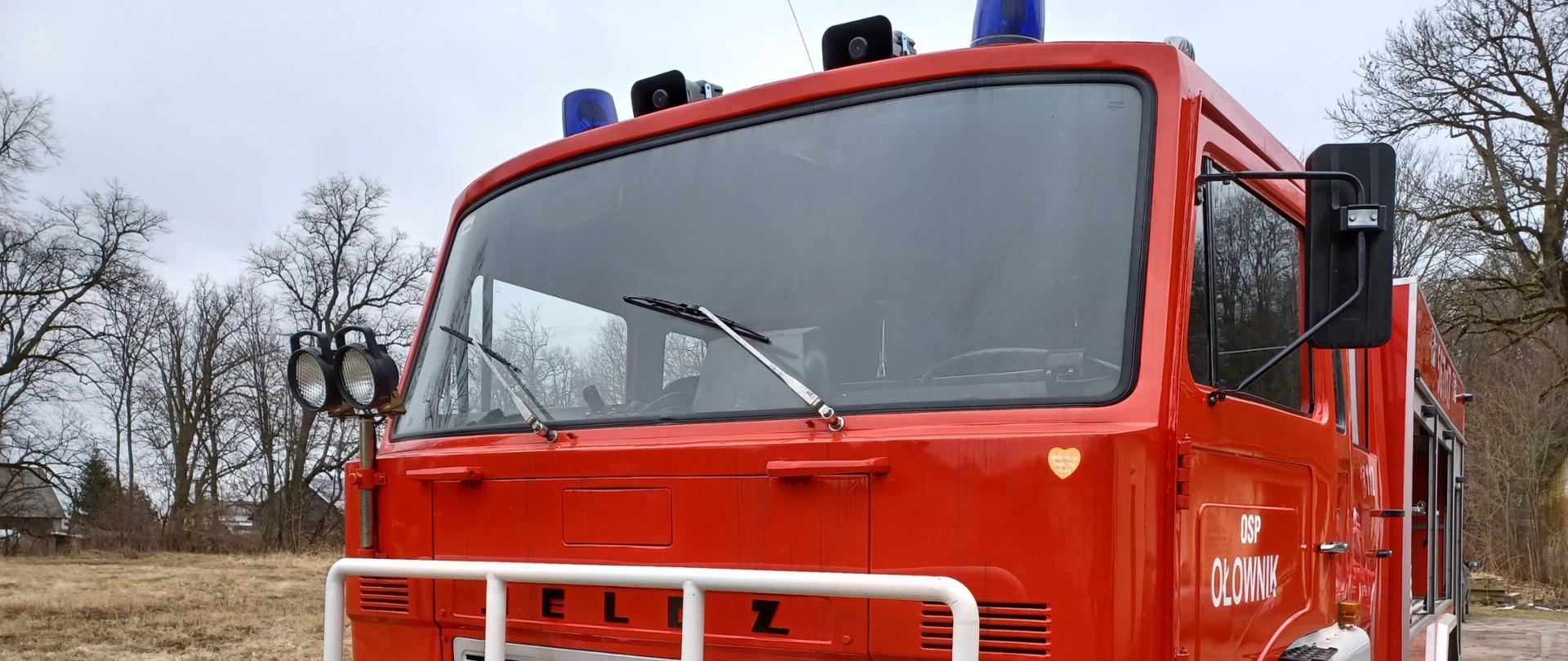 Samochód ratwowniczo-gaśniczy marki JELCZ stoi na podwórku. W oddali za nim widać drzewa i domy. 