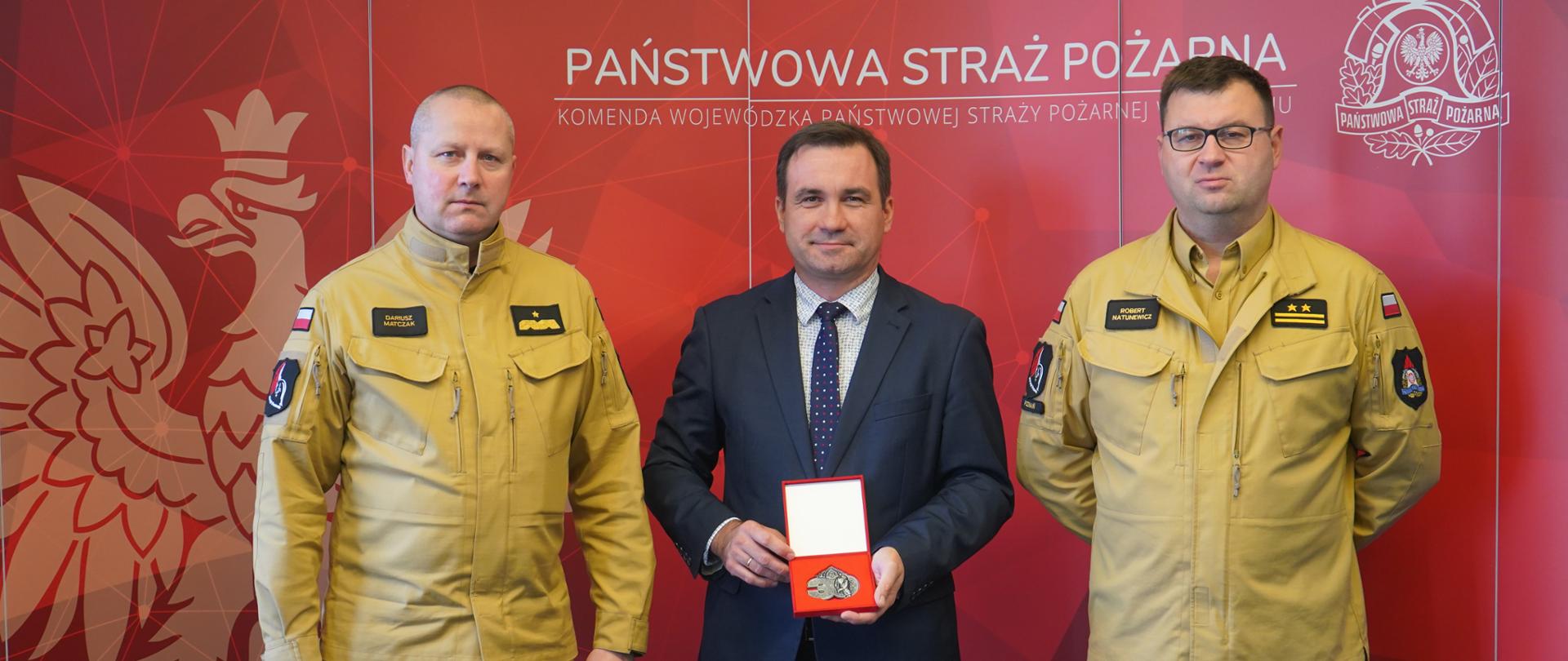 trzech mężczyzn stoi na tle czerwonego baneru, w środku mężczyzna w garniturze trzyma medla , obok to strazacy w jasnych mundurach