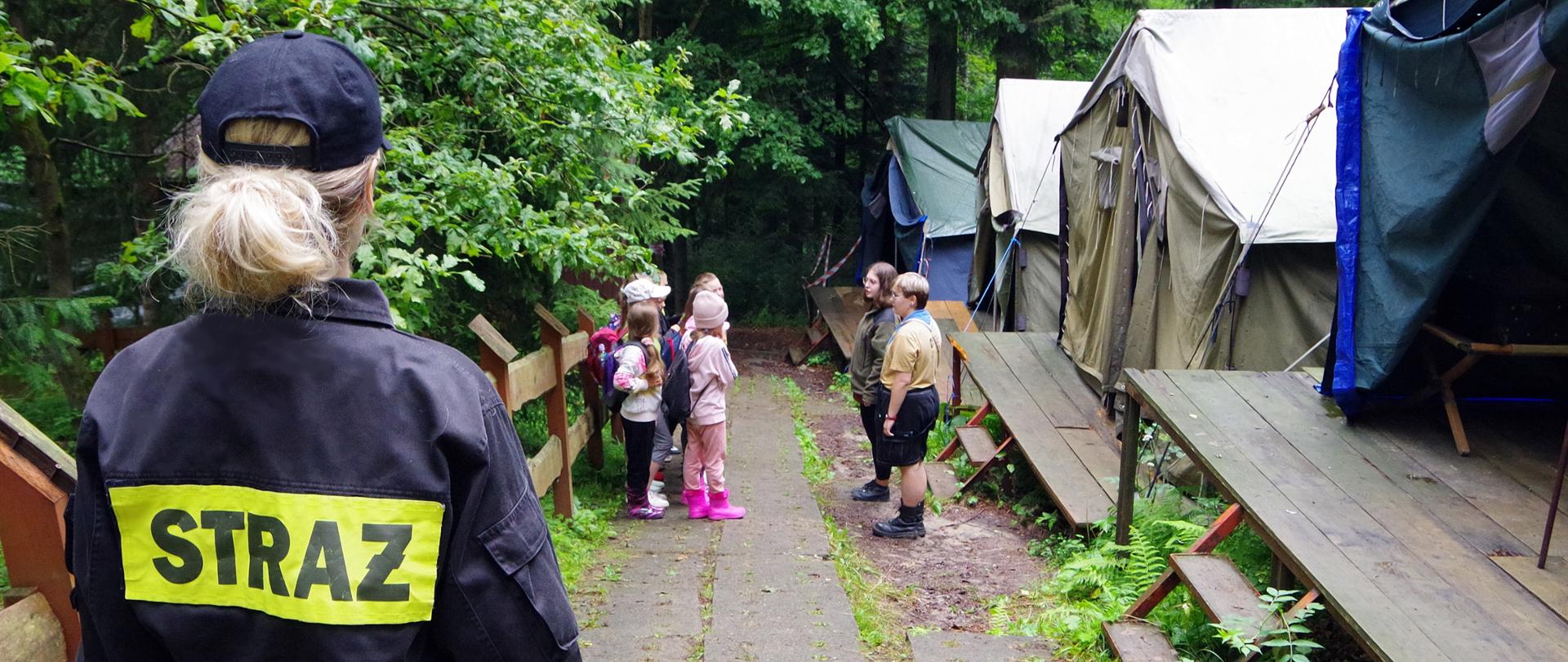 Na pierwszym planie po lewej stronie stoi funkcjonariuszka PSP (widoczna od tyłu). Na drugim planie, bokiem, pododdział harcerzy w dwuszeregu. Po prawej stronie widoczne namioty.