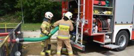Dwóch strażaków ubranych w ubrania specjalne i hełmy podłącza wąż ssawny pomiędzy zbiornikiem przeciwpożarowym, a samochodem pożarniczym. Obok strażaków, na ziemi leży odcinek. Dwie skrytki pojazdu są otwarte.