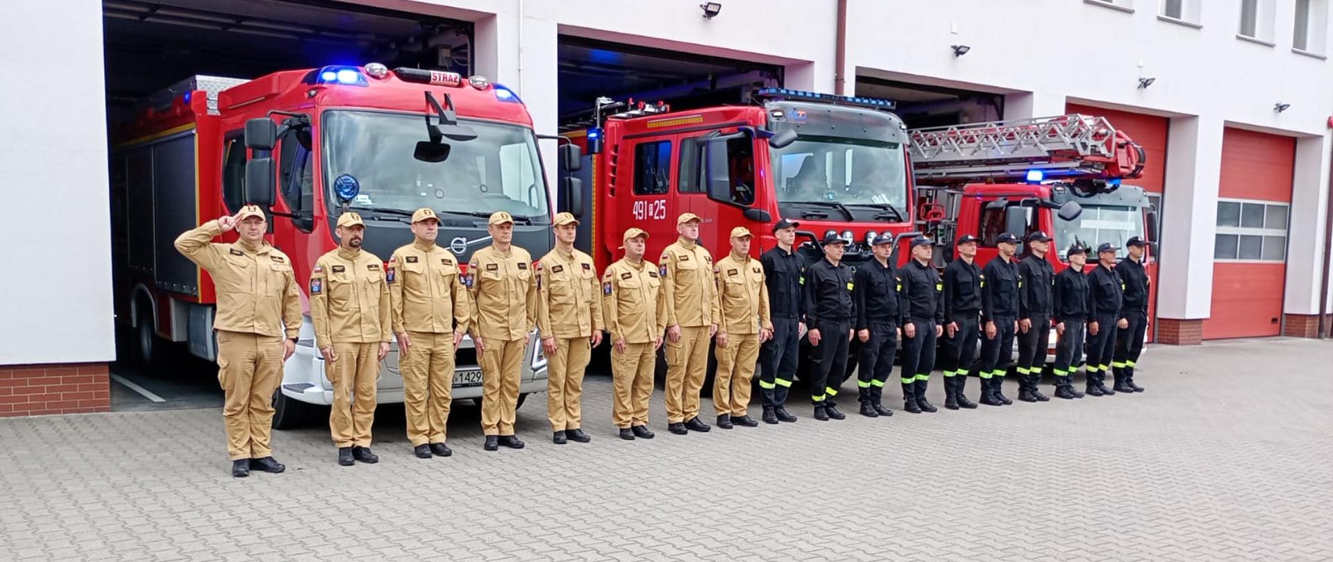 Zdjęcie przedstawia strażaków podczas oddawania hołdu poległym strażakom z poznańskich jednostek ratowniczo gaśniczych. Strażacy stoją na placu przed samochodami bojowymi, które mają włączone światła alarmowe koloru niebieskiego.