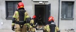 Strażacy PSP ubrani w ubrania specjalne, aparaty powietrzne wraz z maskami i w czerwonych hełmach wchodzą do zadymionych wnętrz budynku firmy. 