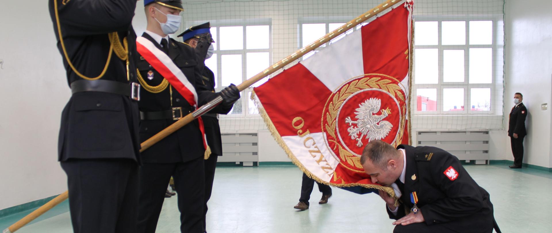 Na zdjęciu widać młodszego brygadiera Wojciecha Pernala, który przyklęknął na czerwonym dywanie przed sztandarem i całuje sztandar. Sztandar Komendy Powiatowej Państwowej Straży Pożarnej w Łęczycy jest pochylony. Na zdjęciu widać strażaków w mundurach wyjściowych. Na twarzach strażacy mają założone białe maseczki. 