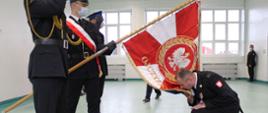 Na zdjęciu widać młodszego brygadiera Wojciecha Pernala, który przyklęknął na czerwonym dywanie przed sztandarem i całuje sztandar. Sztandar Komendy Powiatowej Państwowej Straży Pożarnej w Łęczycy jest pochylony. Na zdjęciu widać strażaków w mundurach wyjściowych. Na twarzach strażacy mają założone białe maseczki. 