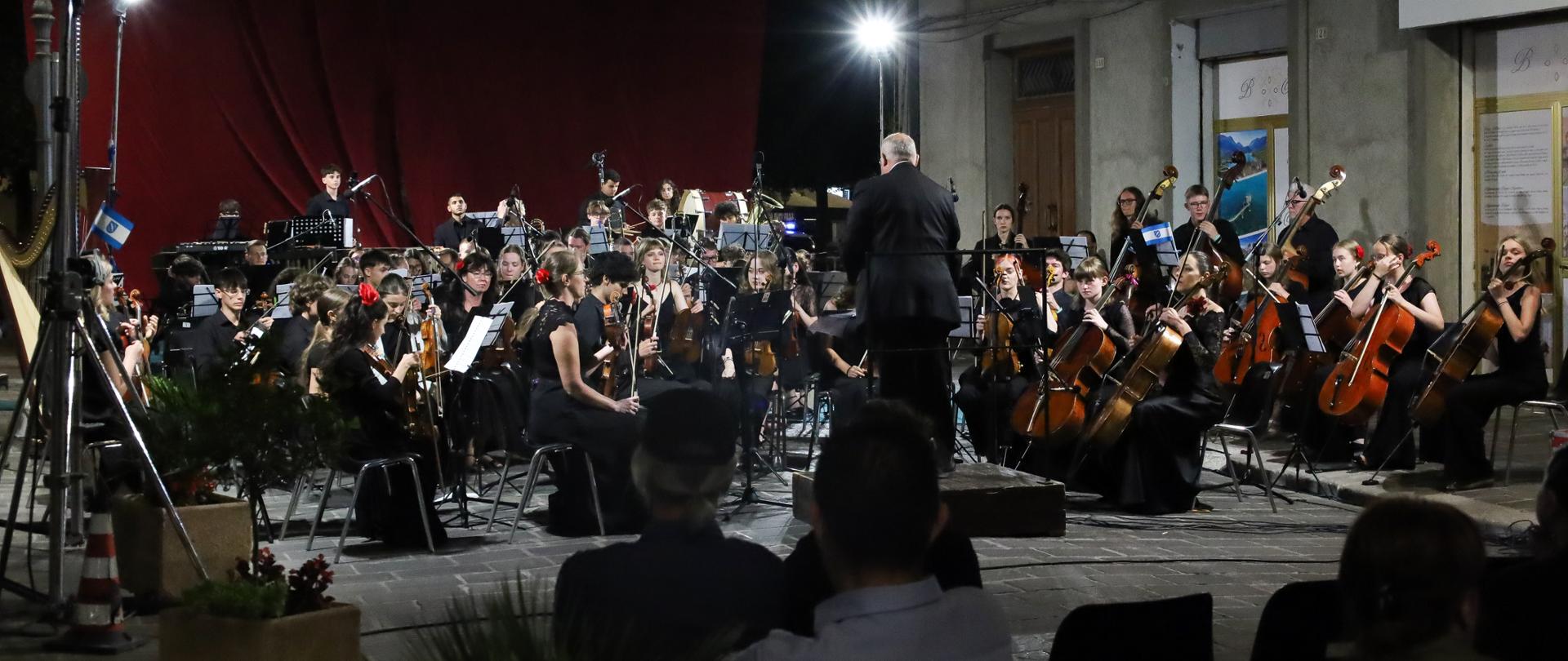 Kolorowe zdjęcie Orchestra Sinfonica Giovanile Europea z koncertu w Atessie w dniu 11.07.2025. Członkowie orkiestry ubrani są w czarne eleganckie stroje i siedzą na krzesłach. Młodzież z Polski ma na sobie czerwone dodatki (dziewczyny czerwone kwiaty we włosach). Przed orkiestrą stoi dyrygent również ubrany na czarno. Orkiestra znajduję się na placu a za nią czerwona kurtyna.