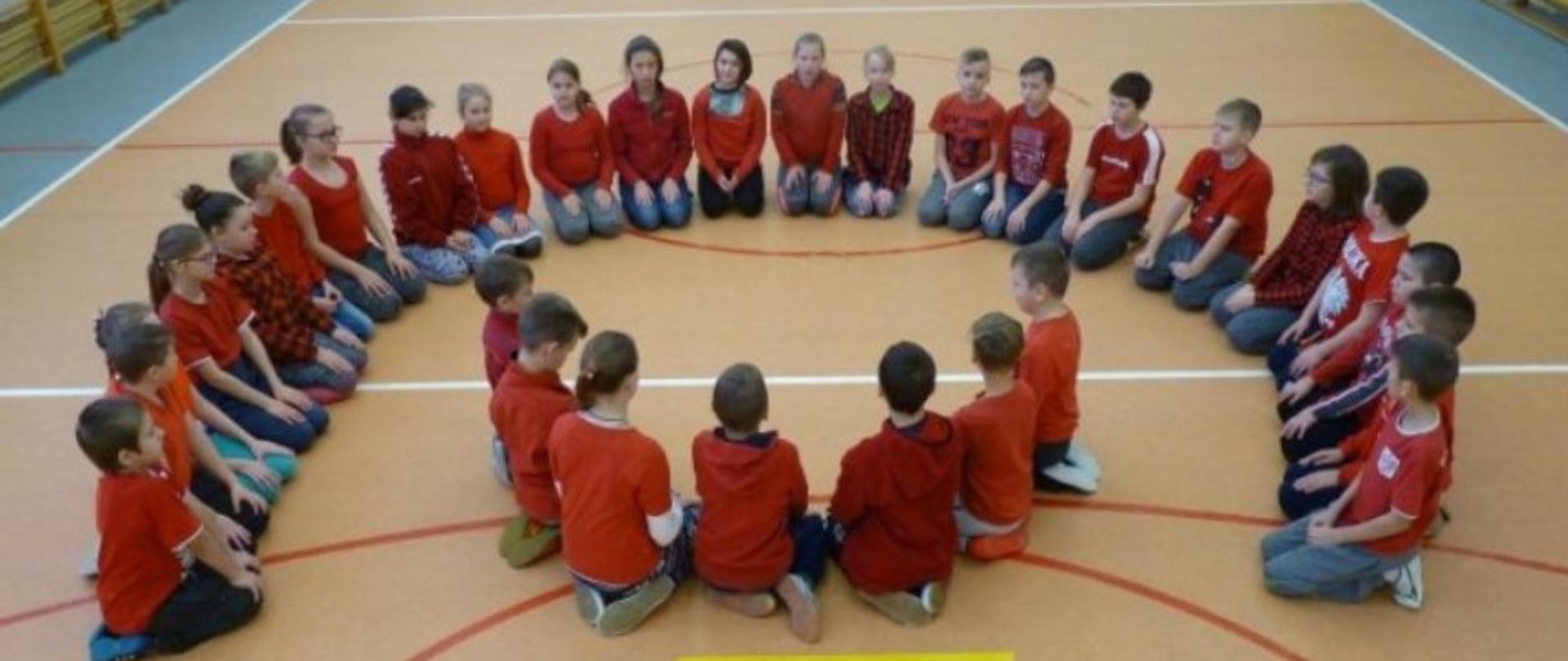 Dzieci w czerwonych koszulkach siedzą na podłodze tworząc kształt logo Polskiej Pomocy. Poniżej jest napis „polska pomoc”.