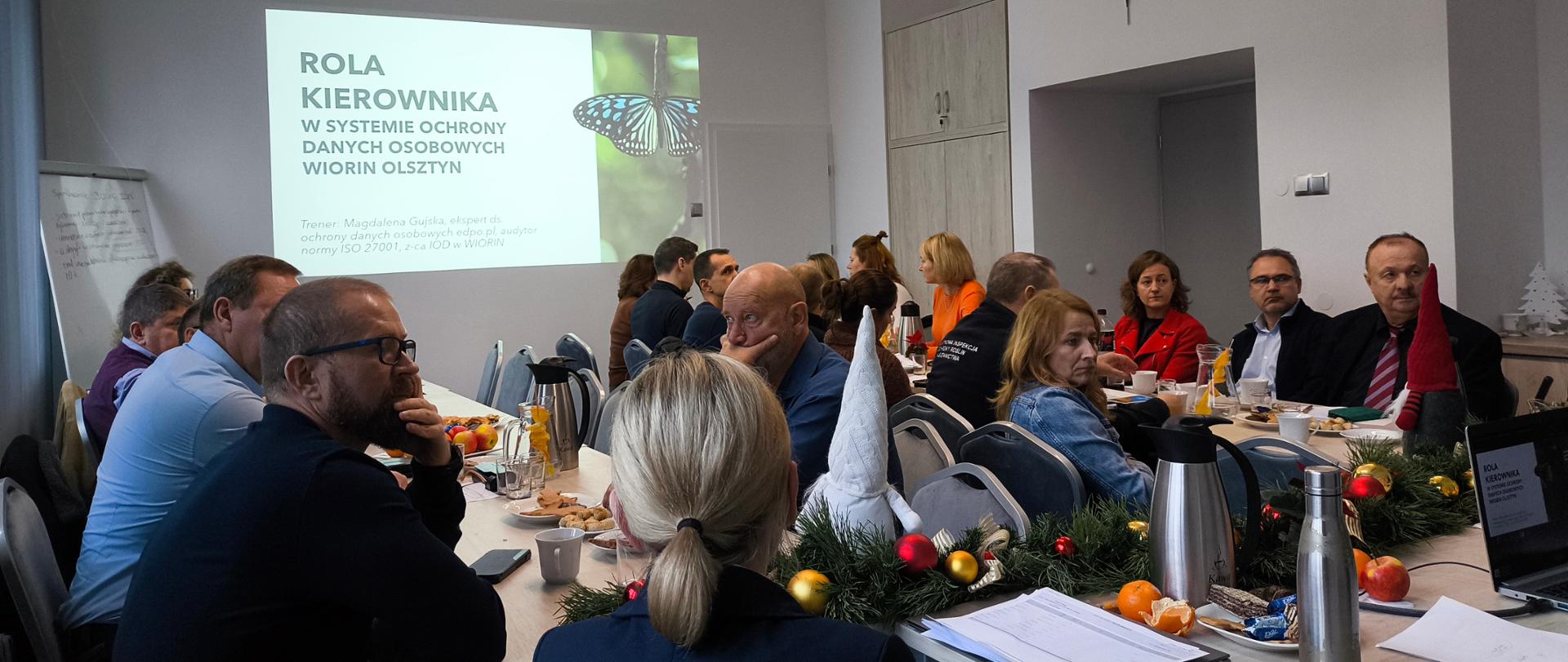 Grupa osób siedzi przy ułożonych równolegle się siebie stołach, w głębi stoi kobieta, za nią na ścianie napis WIORiN w Olsztynie