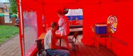 Wewnątrz czerwonego namiotu kobieta w białym kitlu przygotowuje szczepionkę. Obok na krześle siedzi mężczyzna w maseczce.