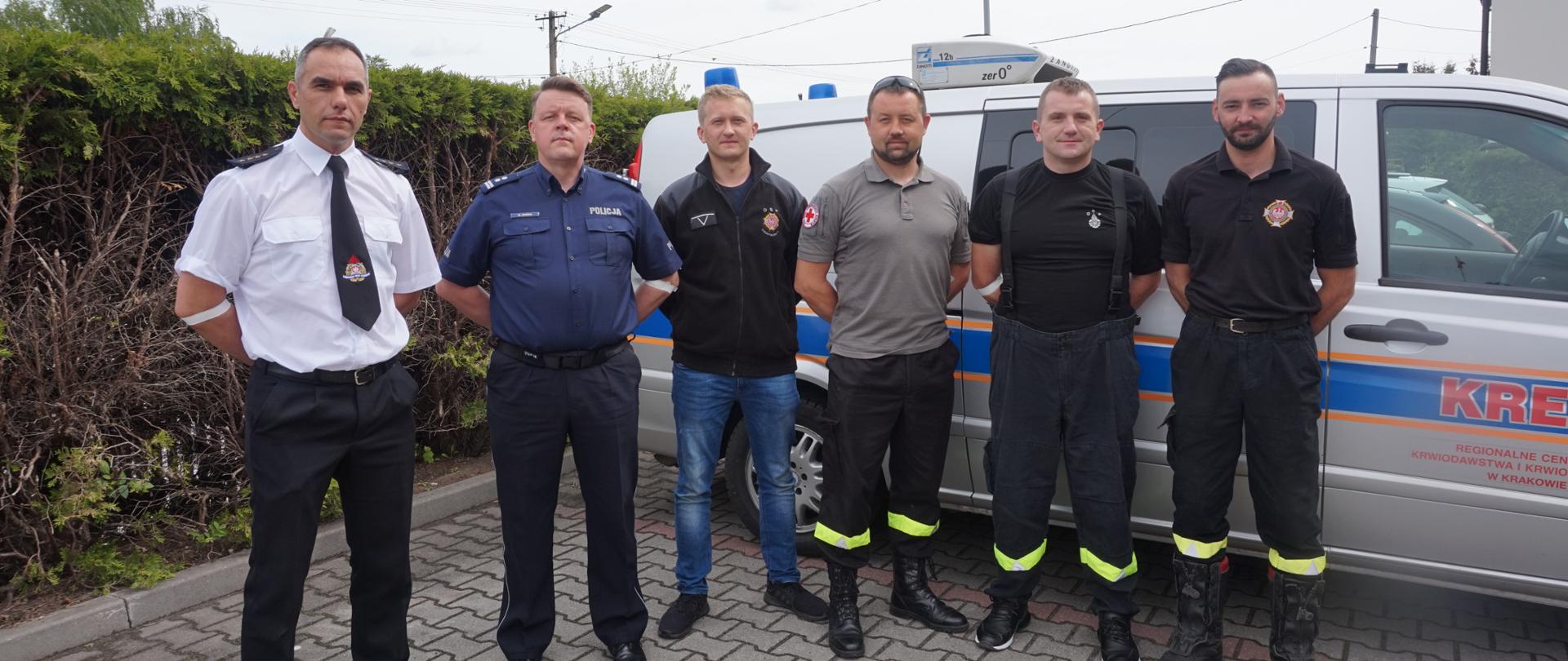 Funkcjonariusze Państwowej Straży Pożarnej i Ochotniczej Straży Pożarnej oraz Policji na tle samochodu Regionalnego Centrum Krwiodawstwa i Krwiolecznictwa na wspólnie pozują do zdjęcia.
