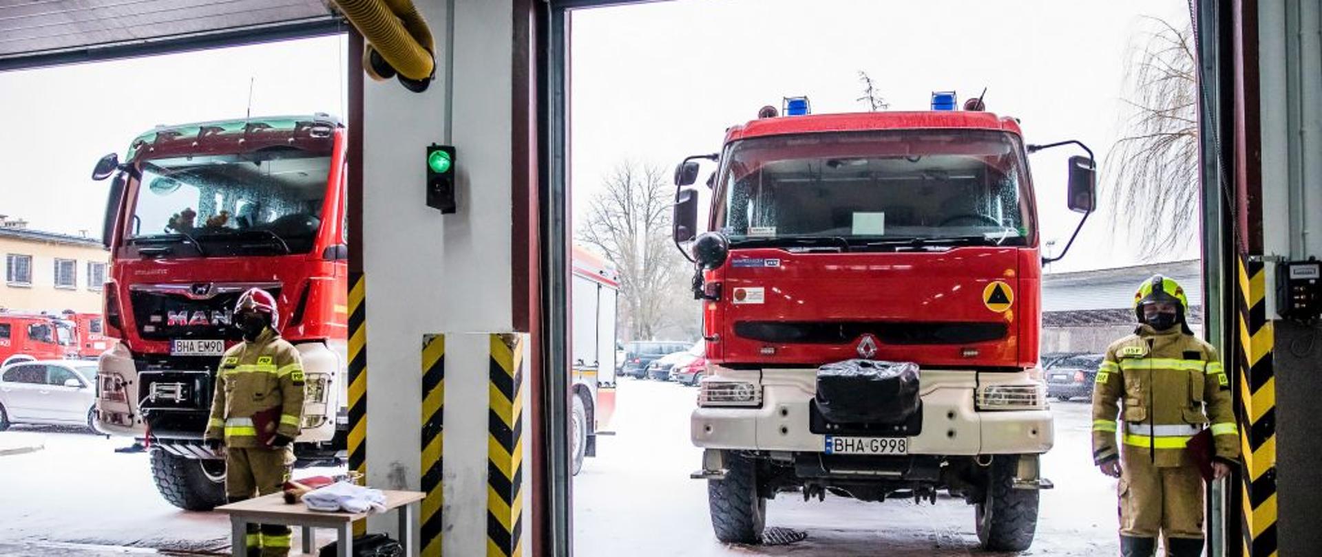 W dwóch bramach garażowych stoją dwa czerwony strażackie wozy bojowe. Przed każdym stoi strażak w żółtym mundurze bojowym.