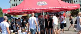 Grupa dzieci i dorosłych stojące przed stoiskiem edukacyjnym PSP, widoczny czerwony namiot stoiska z napisem Komenda Powiatowa Państwowej Straży Pożarnej w Pleszewie