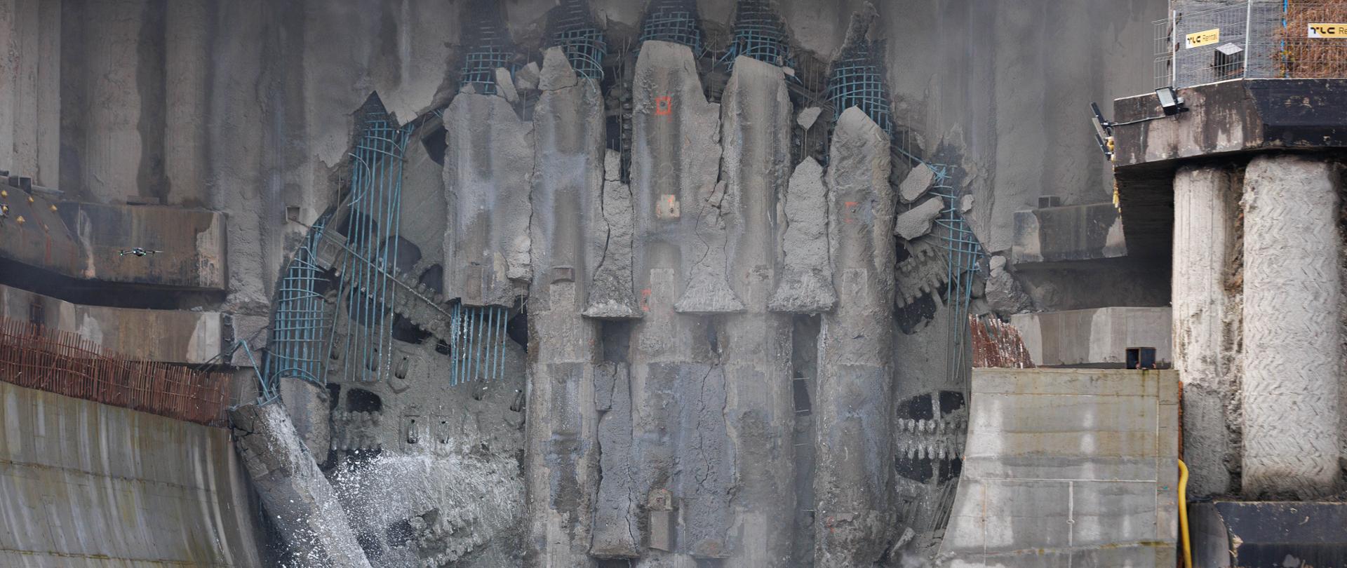 Zdjęcie pokazuje wylot tunelu w trakcie przebicia. W centrum znajduje się duża, półokrągła ściana z kruszącym się betonem i wystającymi prętami zbrojeniowymi. W dolnej części urobek i odłamki zsuwają się na ziemię. Po bokach widać masywne betonowe elementy konstrukcyjne oraz fragment żółtego sprzętu budowlanego.