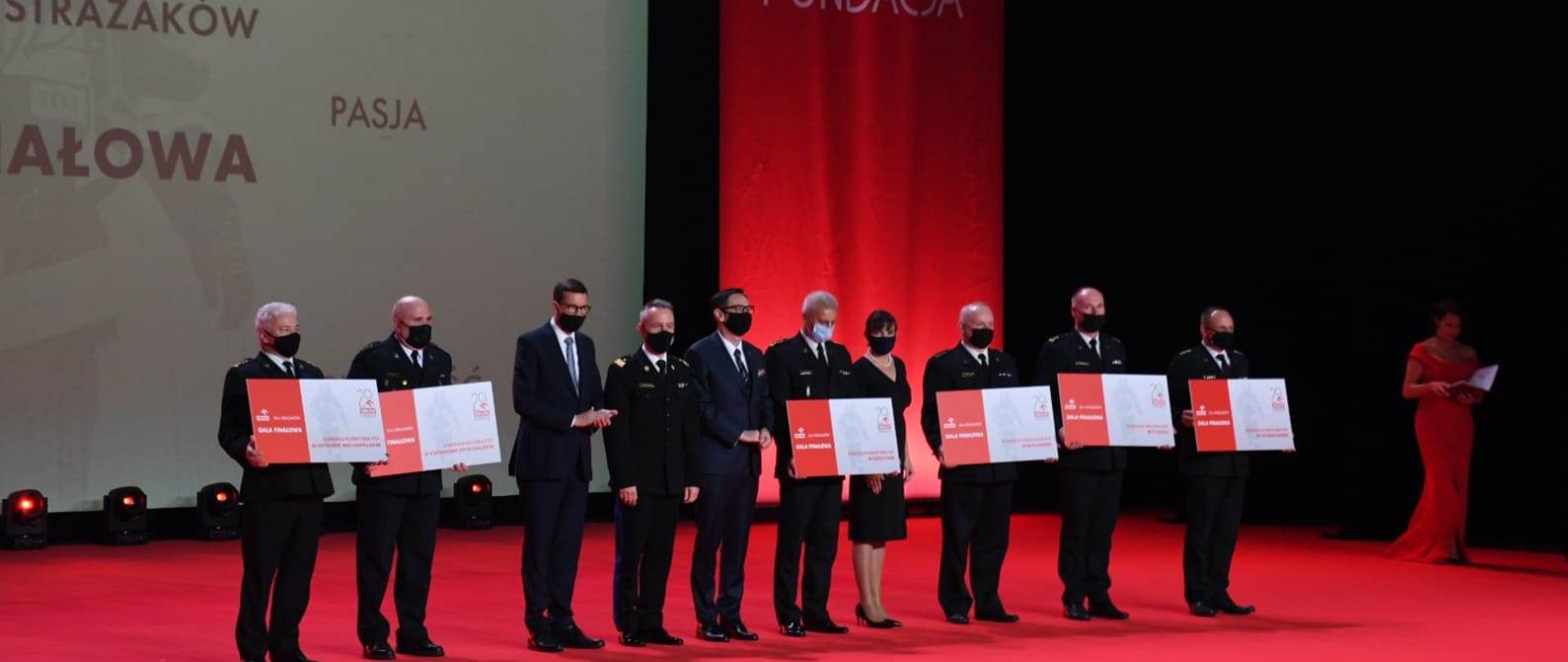 Na scenie 11 uczestników gali, w tym premier, komendant główny , prezes PKN ORLEN, 6 osób trzyma symboliczne czeki