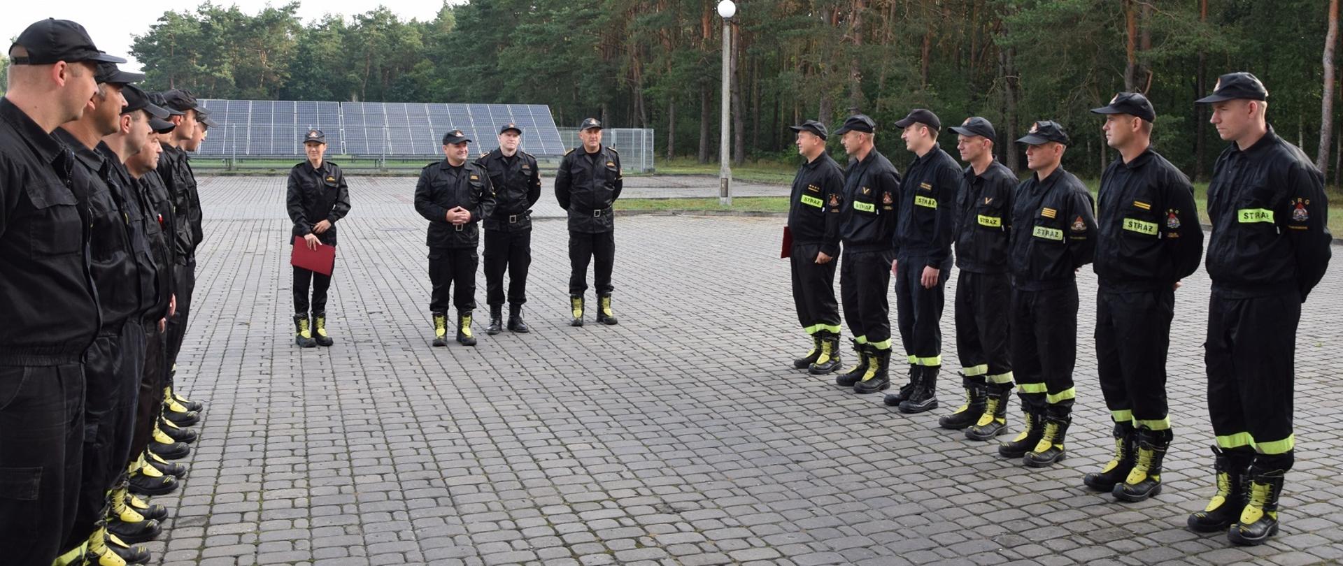 Strażacy w umundurowaniu koszarowym w kolorze czarnym stoją ustawieni w dwóch szeregach naprzeciwko siebie, przed nimi stoi czterech strażaków w umundurowaniu dowódczo-sztabowym. Kobieta strażak trzyma w prawej dłoni bordową teczkę.