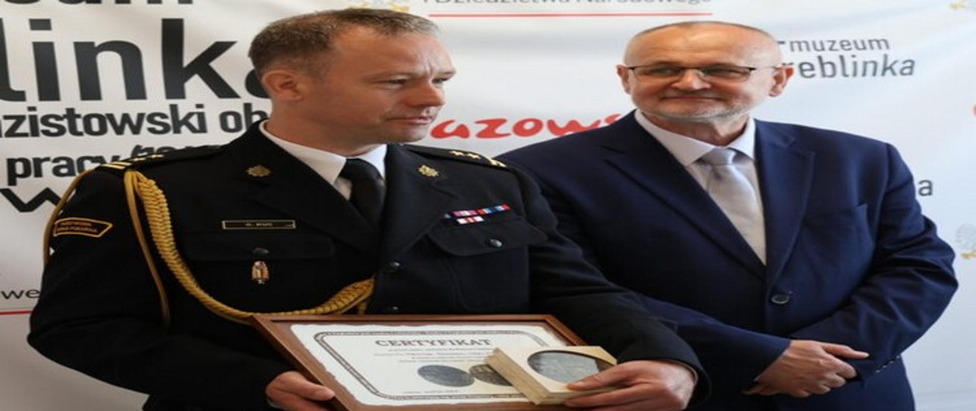 Na zdjęciu widzimy zastępcę komendanta Państwowej Straży pożarnej wraz z otrzymanym medalem oraz dyrektora muzeum w Treblince na tle białego baneru z logo Muzeum Treblinka