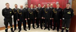 Zdjęcie przedstawia odchodzącego funkcjonariusza w towarzystwie kolegów, strażaków na tle czerwonej ścianki z napisem Państwowa Straż Pożarna