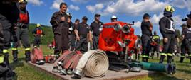 Na zdjęciu widoczni strażacy ochotnicy w ubraniach koszarowych podczas XI Powiatowych Zawodów Sportowo - Pożarniczych na stadionie w Uściu Gorlickim. Przed strażakami na podeście drewnianym pompa pożarnicza koloru czerwonego i węże pożarnicze.