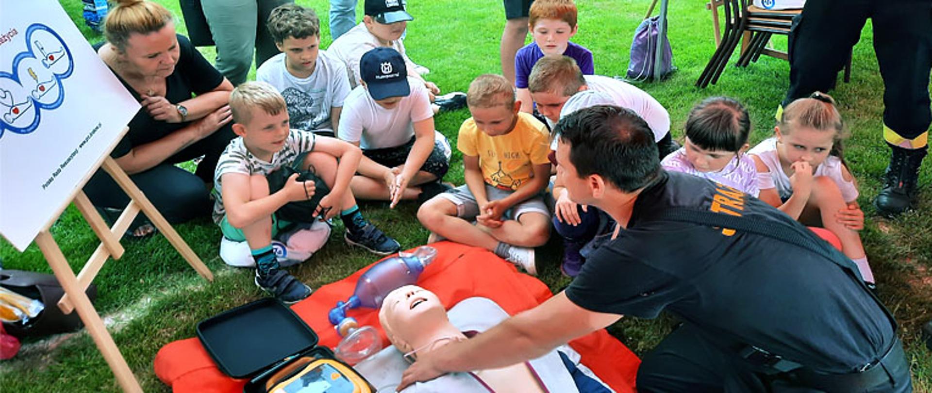 Zdjęcie przedstawia strażaka, który objaśnia przedszkolakom zasady udzielania pierwszej pomocy. Na trawie leży „sztuczny człowiek” typu Phantom do nauki resuscytacji, dookoła niego siedzą dzieci słuchające strażaka, przy dzieciach kuca mama jednego z nich, po lewej stronie zdjęcia stoi na statywie plakat mówiący o zasadach udzielania pierwszej pomocy.