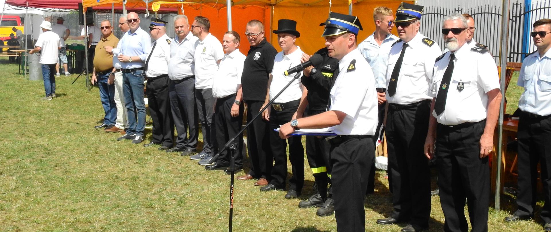 Zdjęcie przedstawia umundurowanych strażaków wraz z współorganizatorami zawodów stojących w szeregu. Na pierwszym planie stoi strażak w białej koszuli mówiący do mikrofonu. W tle widoczne są żółte namioty z napisem Gmina Radzymin.