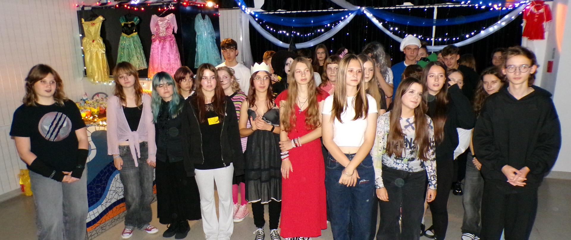 Grupa kilkudziesięciu młodych ludzi w kolorowych ubraniach stoi obok siebie i patrzy w jedną stronę. W tle za nimi zwisają białe i niebieskie pasy oraz łańcuchy choinkowe. Po lewej stronie z sufitu zwisają niebieskie bibułowe kwiaty, a na ścianie wiszą cztery kolorowe sukieneczki.