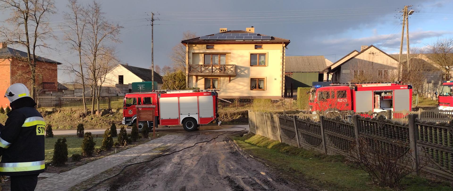 Na zdjęciu widać strażaka stojącego na posesji prywatnej. Na drugim
planie stoją na jezdni przed posesją 3 samochody ratowniczo - gaśnicze.
Za nimi widać domy jednorodzinne.
