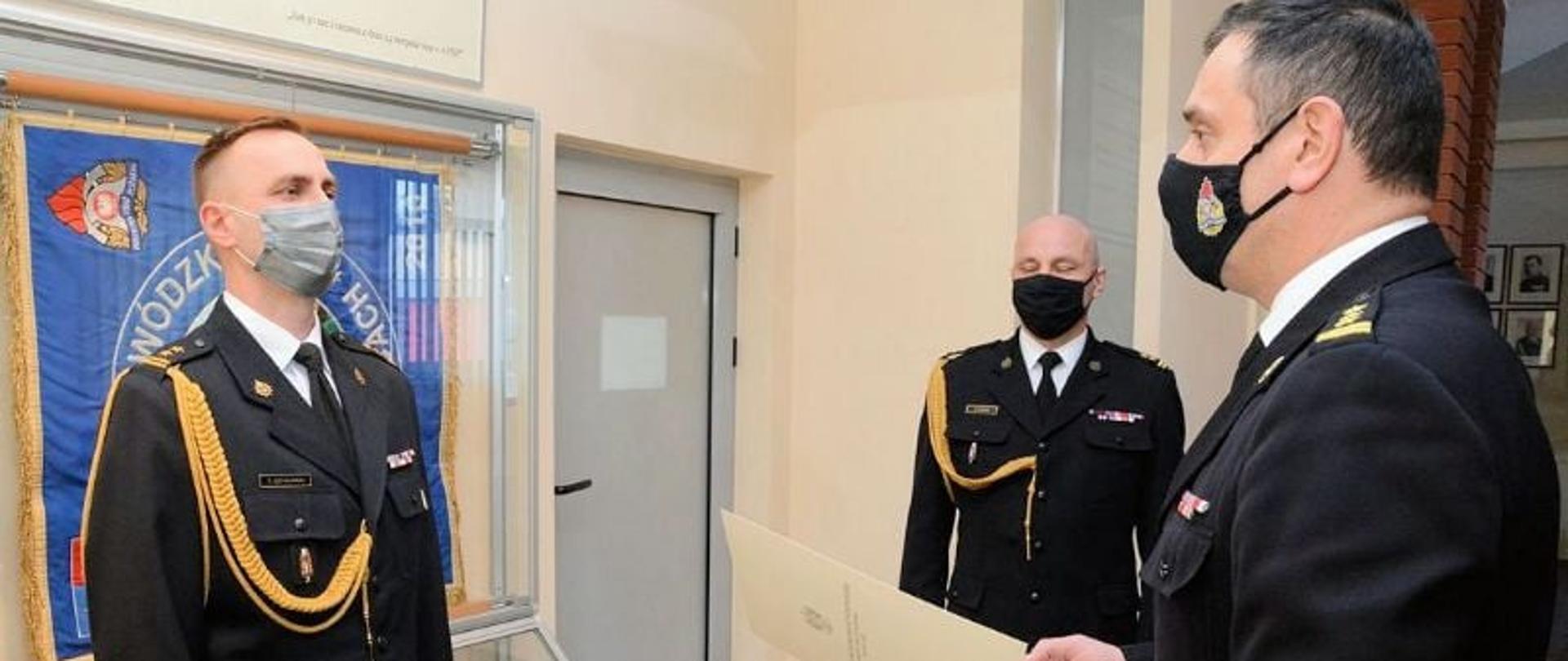 Zdjęcie przedstawia trzech strażaków w mundurach galowych. Ich twarze zasłonięte są maseczkami ochronnymi. Jeden z nich (Świętokrzyski Komendant Wojewódzki Państwowej Straży Pożarnej w Kielcach st. bryg. Krzysztof Ciosek, znajdujący się na pierwszym planie) wręcza akt powołania na stanowisko Zastępcy Komendanta Powiatowego PSP w Jędrzejowie bryg. Tomaszowi Szymańskiemu. Strażacy znajdują się w pomieszczeniu sali tradycji Komendy Wojewódzkiej PSP w Kielcach.