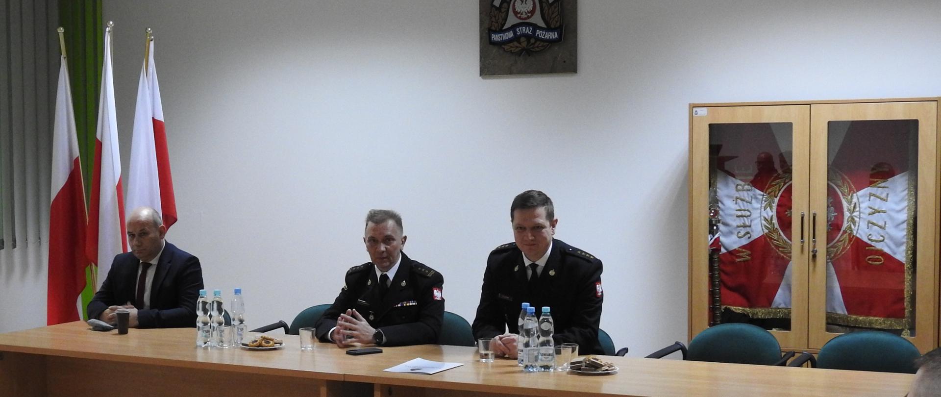 Na zdjęciu znajduje się kilka osób. Na pierwszym planie widać czterech mężczyzn - dwóch strażaków, mężczyznę w mundurze leśniczego oraz mężczyznę w garniturze. W lewej części na drugim planie znajdują się flagi Polski. W prawej części zdjęcia w szafce znajduje się sztandar. Za dwoma mężczyznami w czarnych mundurach, na ścianie znajduje się znak Państwowej Straży Pożarnej.