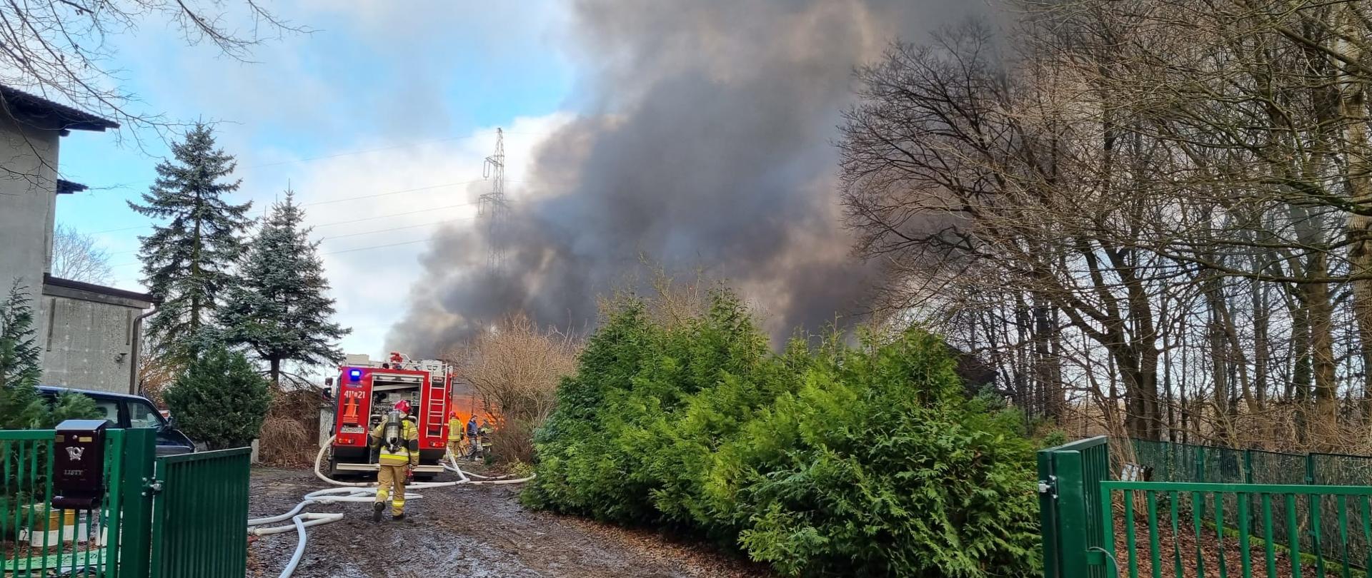 W centralnym miejscu kartu w oddali unoszące się kłęby dymu na pożarem. W około ogrodzenie posesji i rosnące krzewy i drzewa. W oddali widziany od tyły wóz strażacki i jeden strażak idący w kierunku pożaru.